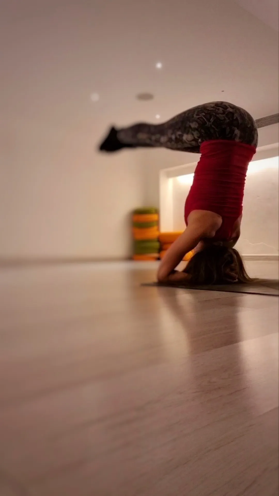 Deena in a supported headstand, legs extended, in a warm-lit studio.