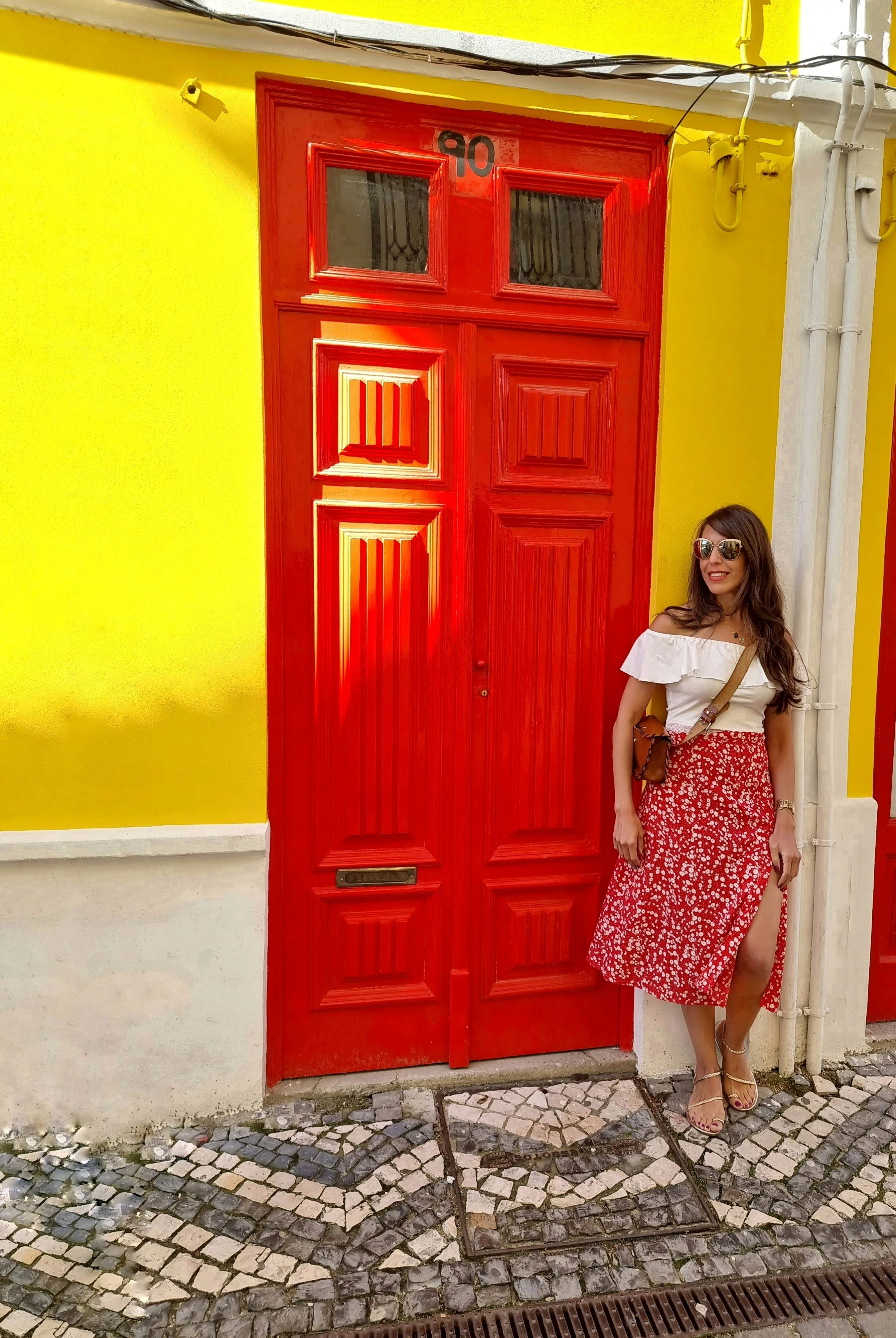 Deena beside a bright red door set into a yellow wall on a cobbled street.